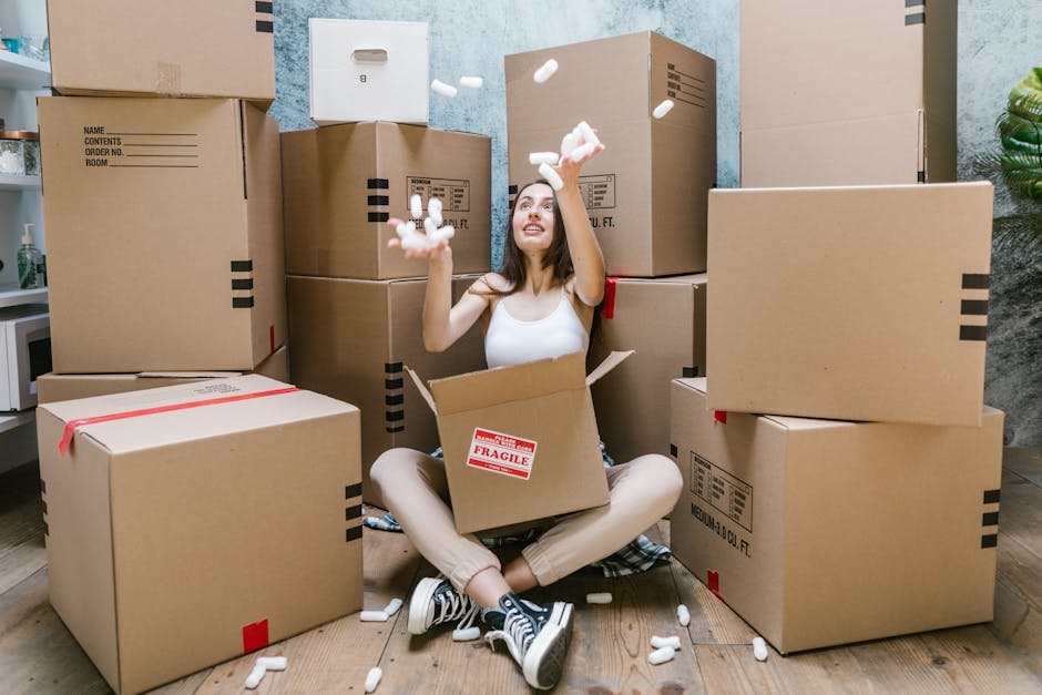 A young woman with long brown hair, dressed in a white tank top and beige trousers, is sitting cross-legged on a wooden floor inside a home, surrounded by numerous large cardboard moving boxes. She is