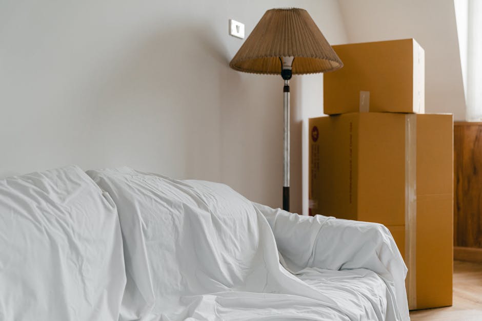 A young woman with long brown hair, dressed in a white tank top and beige trousers, is sitting cross-legged on a wooden floor inside a home, surrounded by numerous large cardboard moving boxes. She is