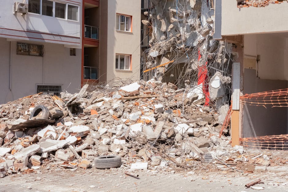 An exterior scene showing a large pile of demolition debris consisting of broken concrete, bricks, twisted metal rods, and scattered construction materials accumulated at the base of a partially demol