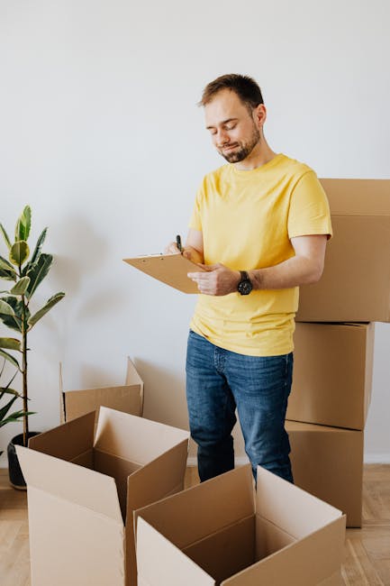 A man with short dark hair and a beard, wearing a yellow t-shirt and blue jeans, is standing inside a room preparing for a home relocation. He is holding a clipboard and a pen, inspecting or making no